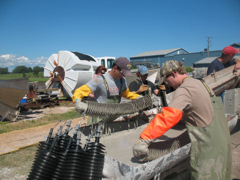 Two young men, wearing ball caps and long gloves, are dipping a series of small round plastic discs into a trough filled with a cement and lime mixture. 