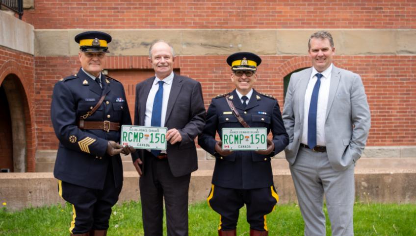 image of four people standing shoulder to shoulder in front of a building and holding a license plate