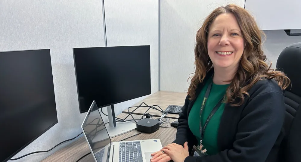 A smiling woman with shoulder-length brown hair sits at a desk.