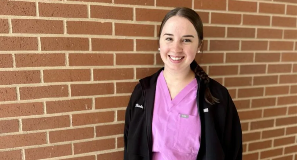 A brunette young woman wearing pink scrubs smiles with a brick wall behind her.