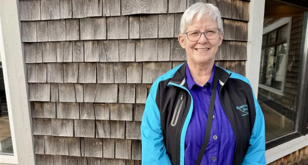 A white woman with short grey hair smiles, with weathered shingles behind her.