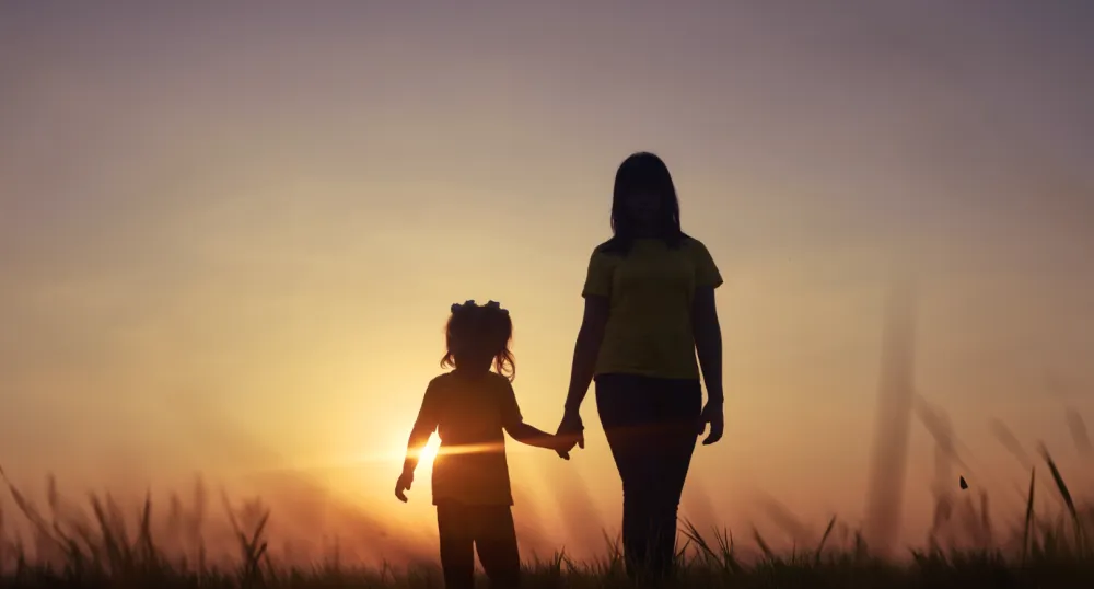 Silhouette of an adult and a child holding hands while walking through a grassy field at sunset, with the sun shining brightly behind them.