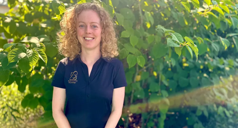 A blonde, curly-haired woman smiles in front of lush green foliage.
