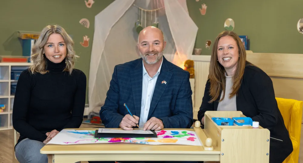 Two women and a man sit at a child's table in a playroom.