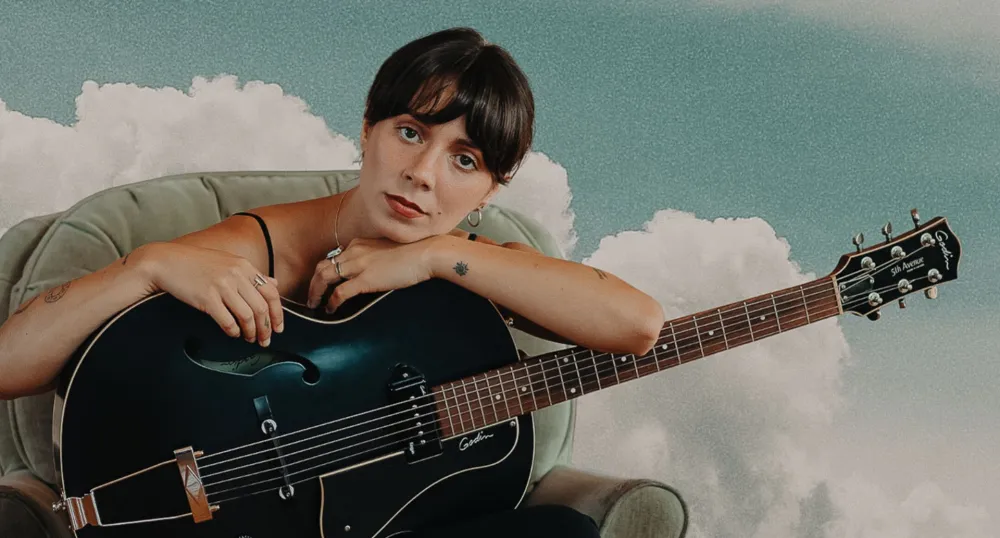 Une femme tient une guitare noire avec un ciel bleu et des nuages derrière elle.