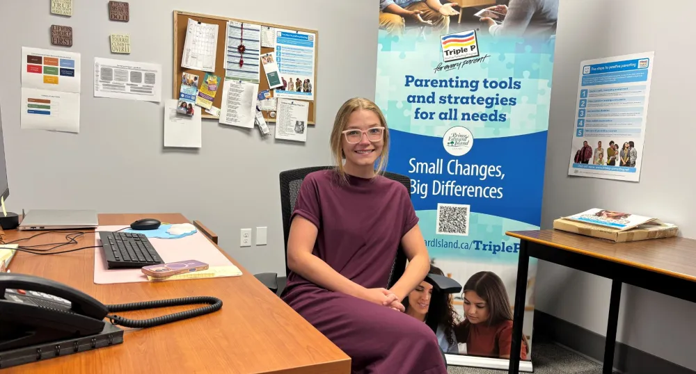Une femme blonde est assise à son bureau avec un poster de Triple P Parenting derrière elle.