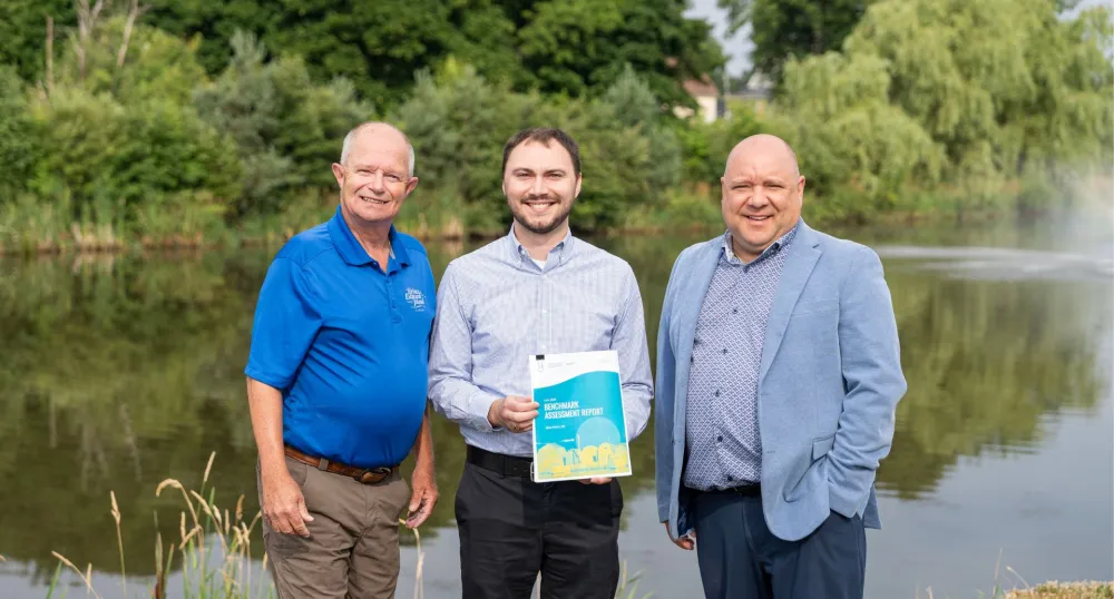Trois hommes blancs se tiennent côte à côte devant un étang avec en arrière-plan des arbres verts.