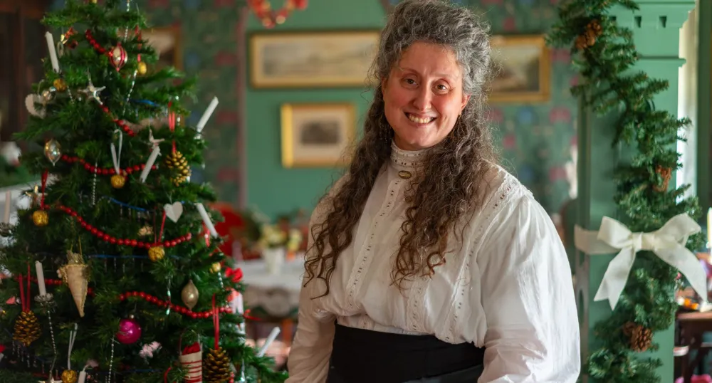 Une femme aux longs cheveux gris sourit devant un sapin de Noël.