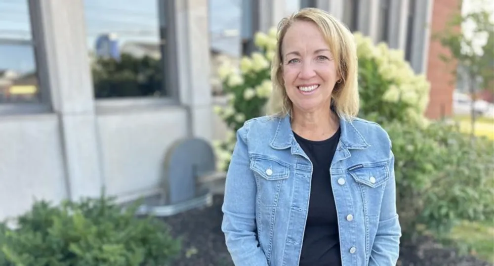 A blonde-haired woman wearing a denim jacket smiles outside a brick building.
