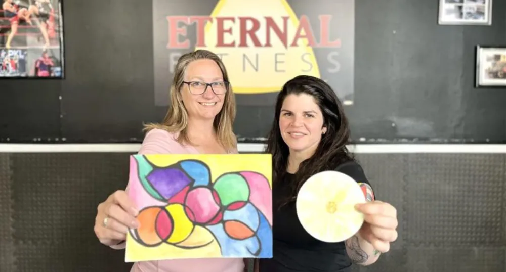A blonde woman and a brunette woman stand next to eachother in front of the Eternal Fitness sign.