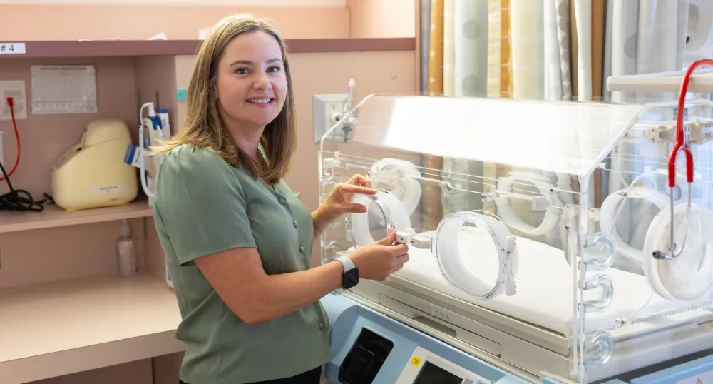 Dr. Maclean with shoulder length hair stands next to a NICU incubator.