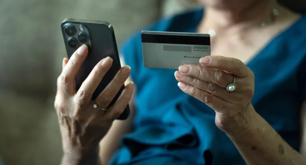 An elderly woman holds a cell phone in one hand, and a credit card in the other.