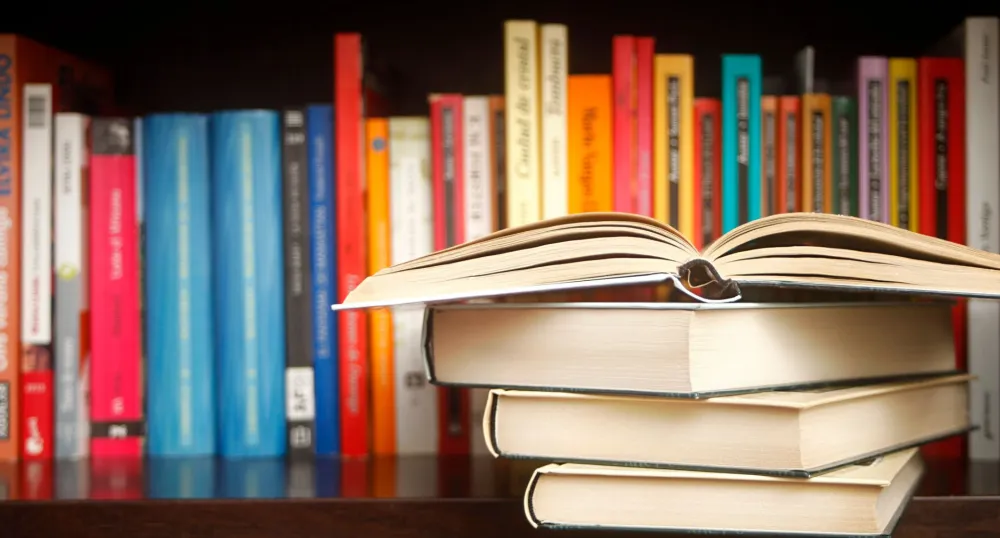 A stack of books set in front of a colourful row of books on a shelf.