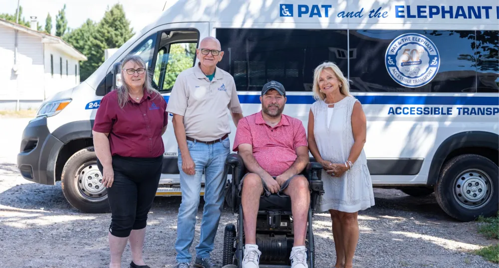 Three people stand around a man in a wheelchair in front of a Pat and the Elephant van.