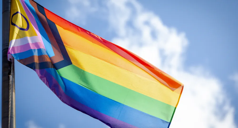 The pride flag blows in the wind with a bright blue sky in the background.