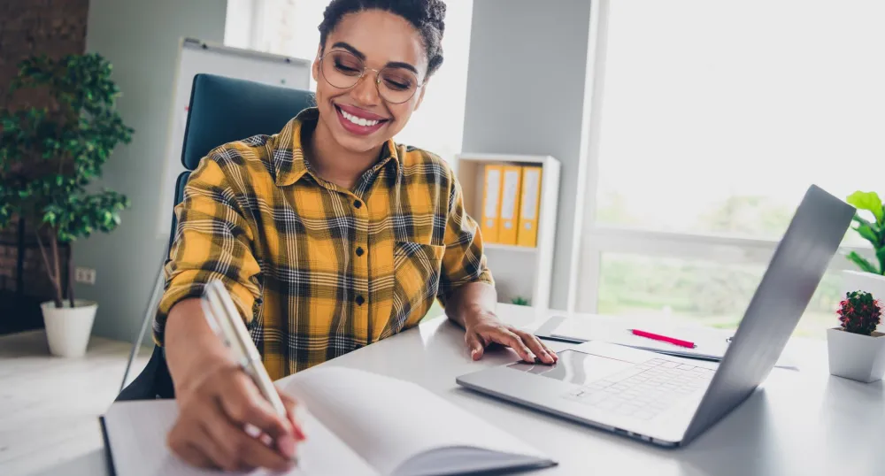 A person sitting at a desk holding a pen to a notebook and their laptop is open.
