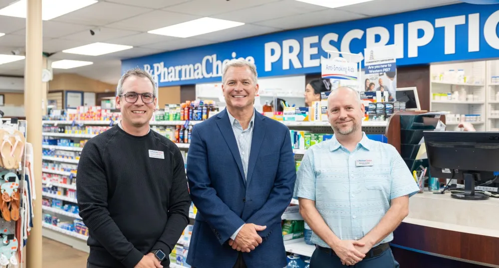 Two male pharmacists and the minister stand side by side in front of a pharmacy counter.