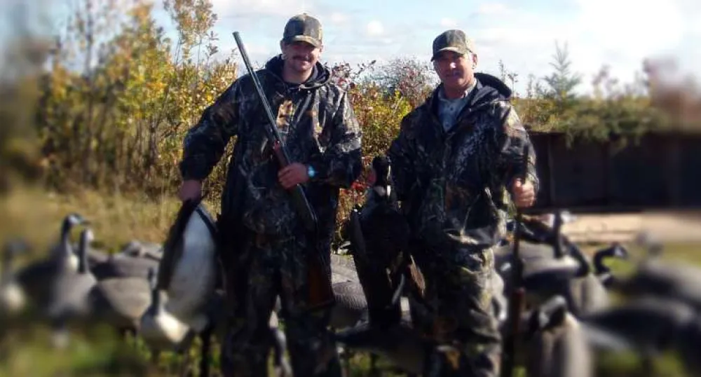 Two men with goose decoys in background. One is holding a dead goose.