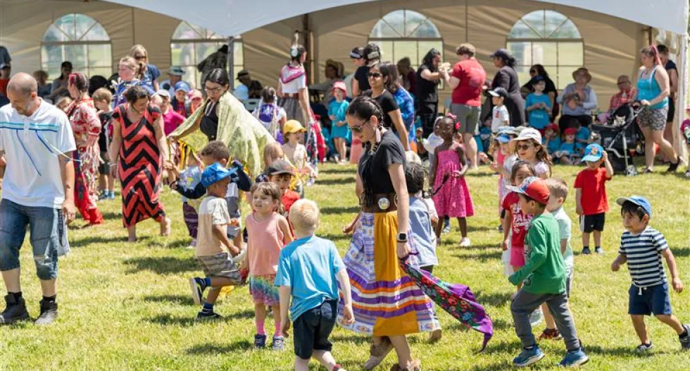 Children and adults gathered together infront of a white tent for a Pow Wow in PEI.