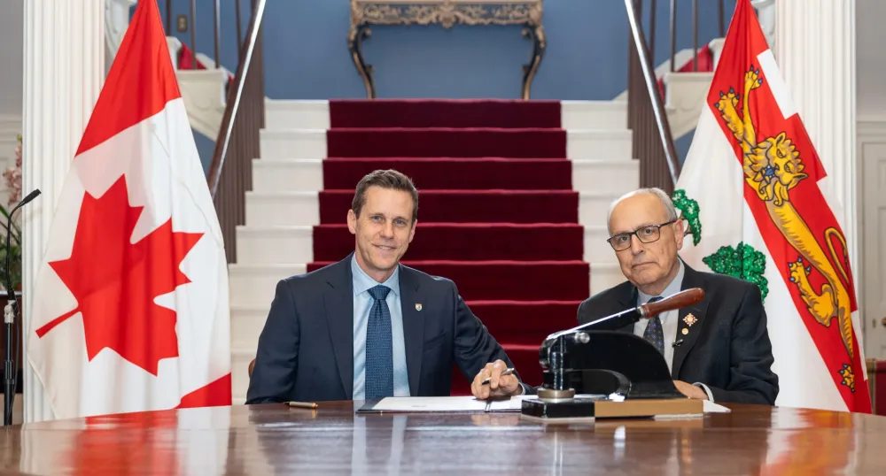 Le premier ministre et le lieutenant-gouverneur assis entre deux drapeaux.