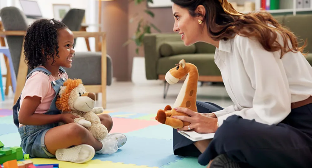 A woman and girl smiling and talking to each other while sitting on the floor holding plush animal toys and a clipboard.