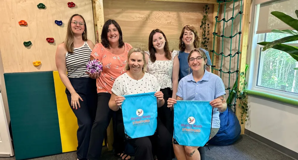 A group of women hold Might Minds bags in front of a rock-climbing wall.