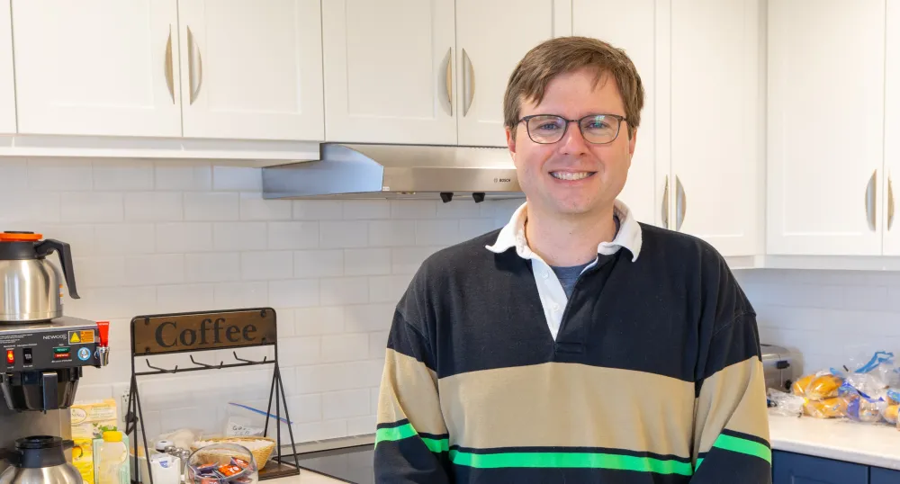 A smiling man wearing glasses and a casual rugby jersey stands in a kitchen