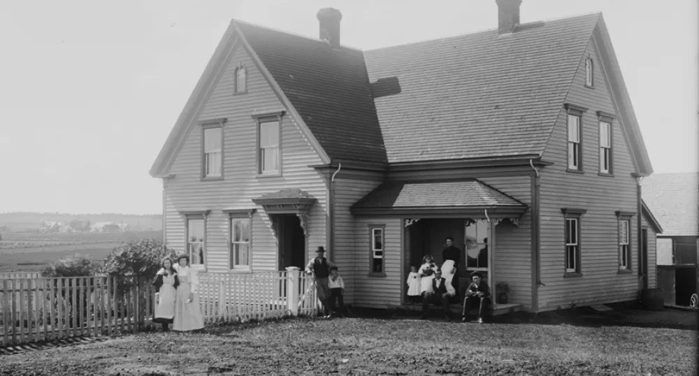 La maison Brehaut à Alexandra, à l’Île-du-Prince-Édouard, vers 1890-1906. On peut voir des membres de la famille prenant la pose sous le porche et devant la clôture, ainsi que le paysage rural en arrière-plan.