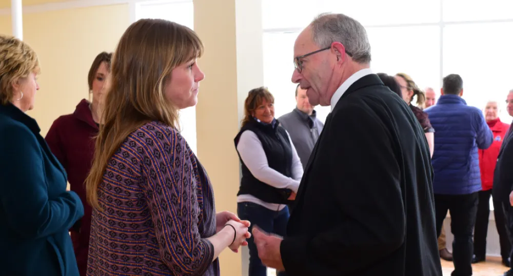Two people facing each other talking while standing in a room filled with people.