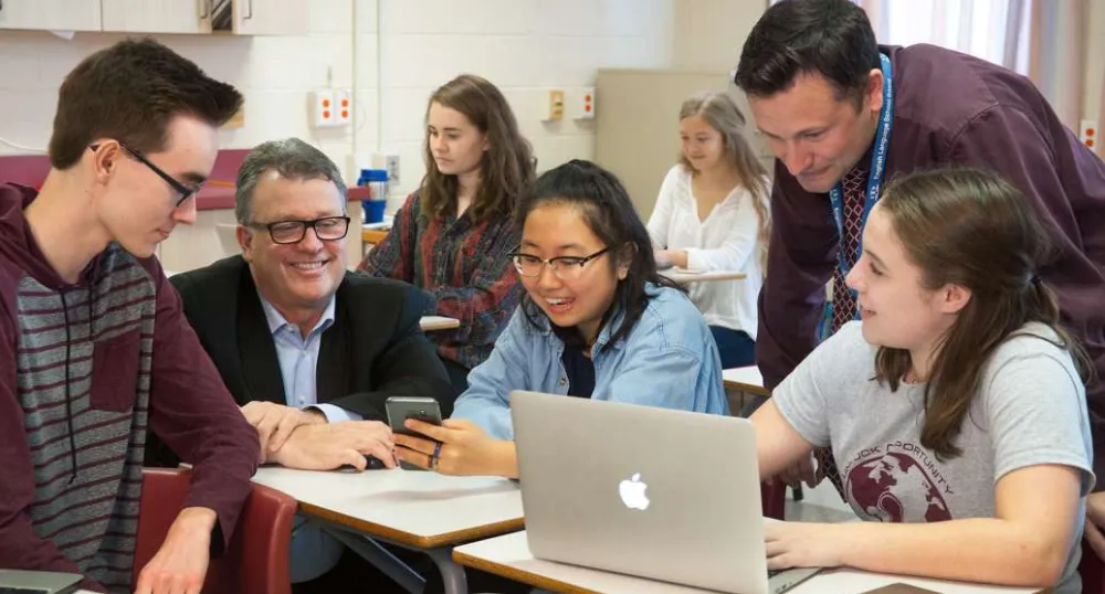 Minister Currie, a teacher and three high school students looking at a smart phone and a cumputer.