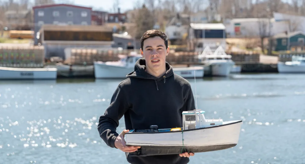 image of a person holding a model fishing boat at a wharf with fishing boats in the background