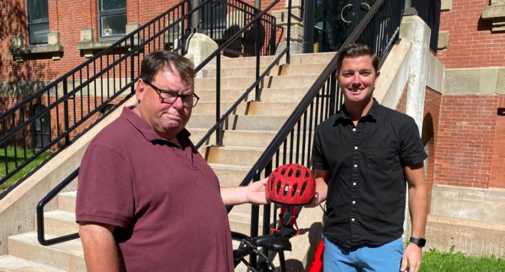 image de deux personnes tenant un casque de vélo avec un vélo en arrière-plan et devant un escalier à l'extérieur d'un bâtiment en brique