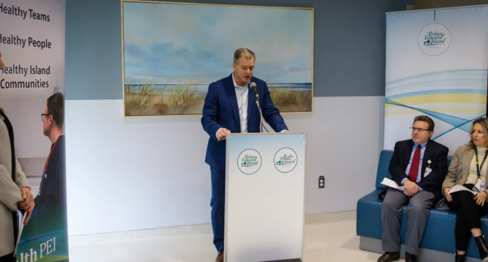 Minister McLane stands at the podium, three people seated to the right look on and one woman on the left is watching. Banners behind him show Island scenery. 