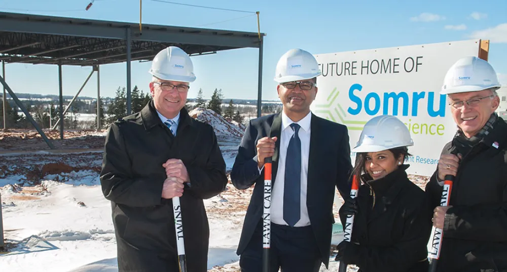 Four people in hardhats holding shovels in front of building under construction
