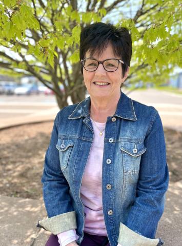 A smiling older woman in a jean jacket sits outside on a sunny day.