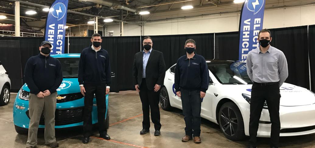 Four Electric Vehicle reps stand in front of two electric vehicles in a convention facility