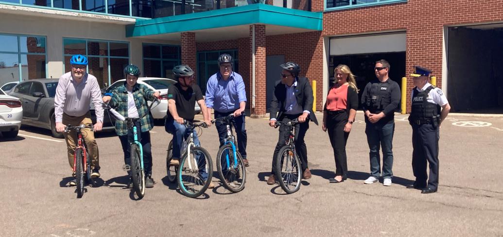 Five people sit upon bicycles in front of the Charlottetown Police Station; three people stand beside them