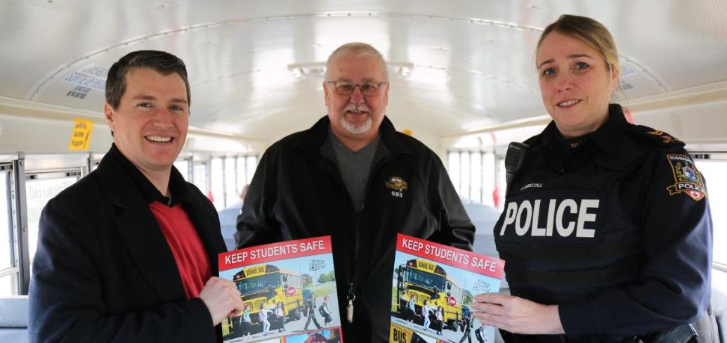 Three people stand inside a school bus holding school bus safety posters