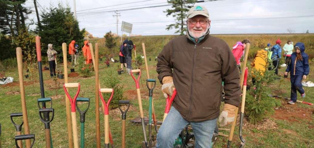 Bill Hogg stands beside a row of shovels with students planting trees in the background