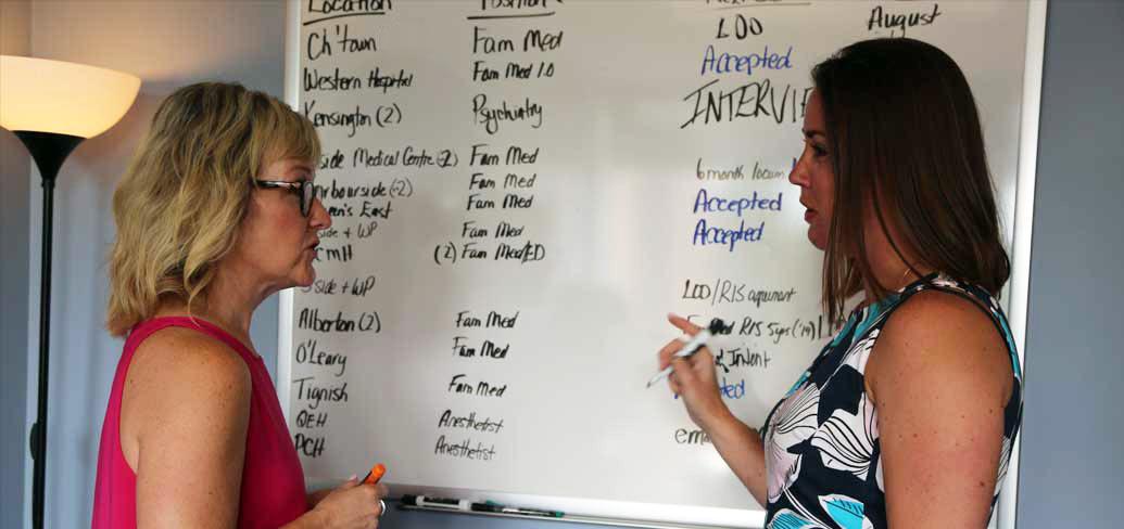 Photo shows two women talking in front of a whiteboard