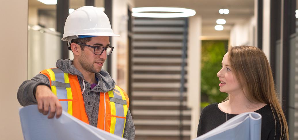 Two people, ne with a hardhat on, holding drafting documents while in a hallays.
