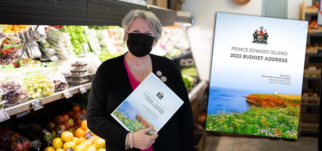 Finance Minister Darlene Compton is standing in a local grocery market holding a copy of the budget