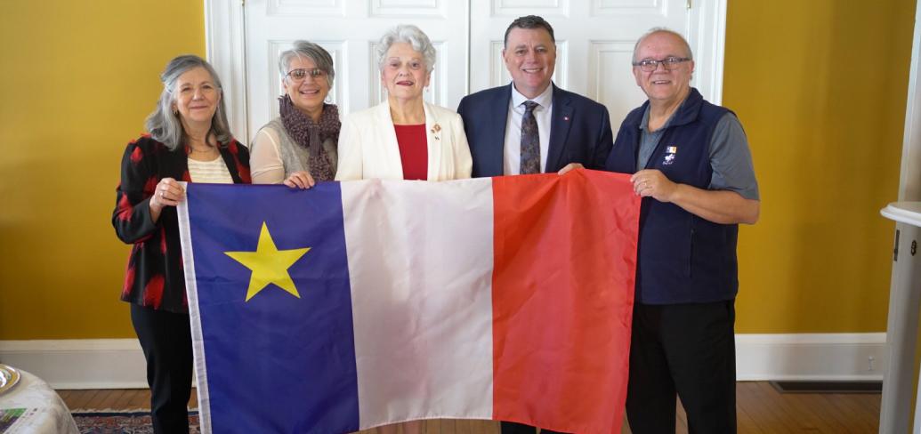  image de cinq personnes debout côte à côte dans une pièce, tenant un grand drapeau acadien.