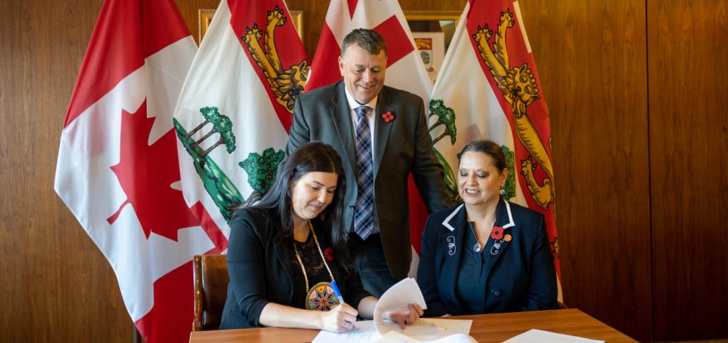 image of two people aitting at a table signing an agreement with another person standing behind them watching with some flags in the background