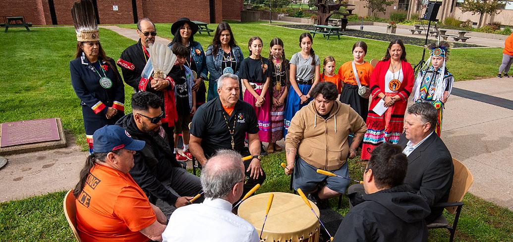 Mi'kmaq drummers, accompanied by Premier King and Mayor Brown sit drumming in a circle outside on a sunny fall day, while dancers look on. 