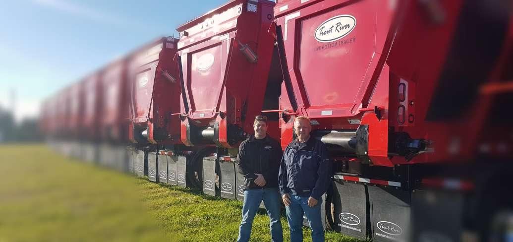 Harvey Stewart and Darrin Mitchell stand in front of a row of red-coloured Trout River Industries live bottom conveyor trailers