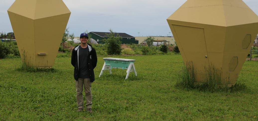 Broyde Chappell stands in a field between two four-metre high hexagon bee hives 