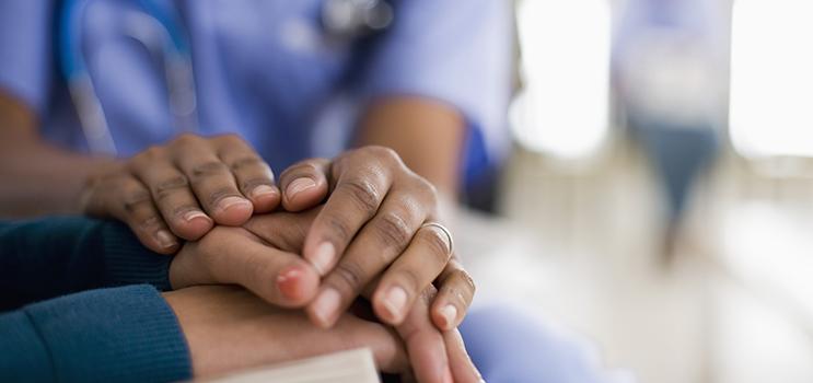 A health professional has their two hands on top of a woman’s hands showing comfort.