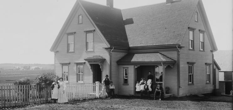 Exterior of the Brehaut house in Alexandra, Prince Edward Island, ca. 1890-1906. The family is posed on the front porch and in front of the fence. Rural landscape in background.