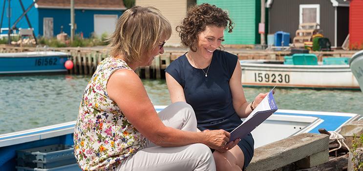 Image of two women sitting on dock at a PEI fishing harbour looking over a brochure